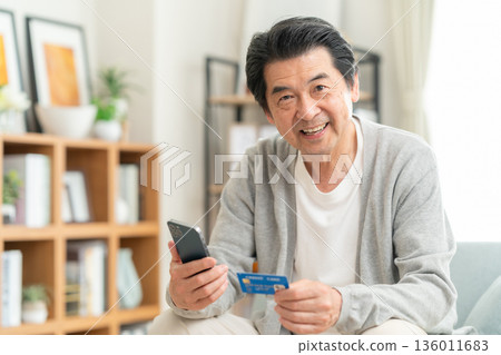 Middle-aged man looking at credit card and smartphone in living room 136011683