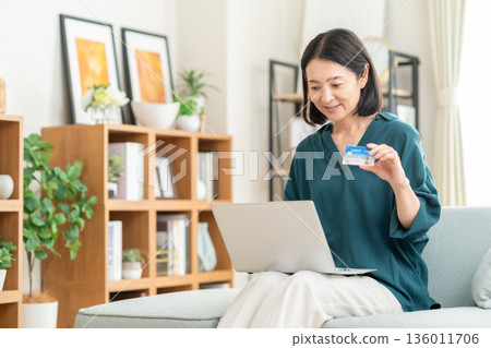 一位中年婦女在客廳看著信用卡和筆記型電腦。 136011706