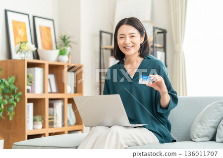 一位中年婦女在客廳看著信用卡和筆記型電腦。 136011707
