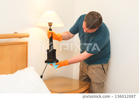 Man in Casual Attire Dusting Under a Desk Lamp, Embracing His Role as a Stay-at-Home Husband in a Tidy and Inviting Living Space Man in Casual Attire Dusting Under a Desk Lamp, Embracing His Role as a Stay-at-Home Husband in a Tidy and Inviting Living Space 136012087