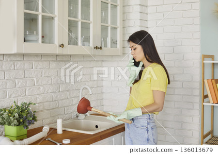 Woman standing in kitchen with plunger talking by mobile phone with plumber trying to unclog a sink Woman standing in kitchen with plunger talking by mobile phone with plumber trying to unclog a sink 136013679