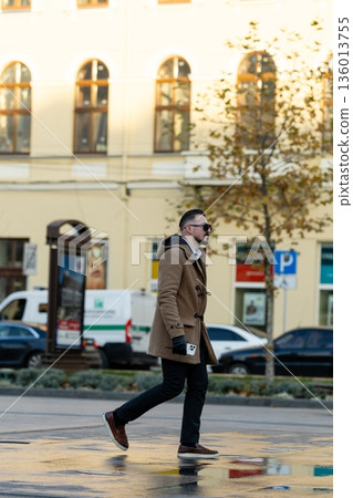 Fashionable man strolling through the vibrant city square on a sunny autumn afternoon, surrounded by historic buildings and parked cars 136013755