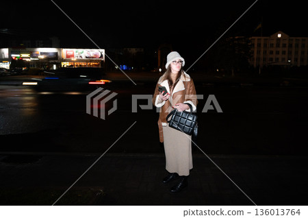 Elegant girl standing on a city street at night, holding a phone and wearing a stylish outfit, with cars passing by and vibrant lights reflecting the bustling urban life Elegant girl standing on a city street at night, holding a phone and wearing a stylish outfit, with cars passing by and vibrant lights reflecting the bustling urban life 136013764