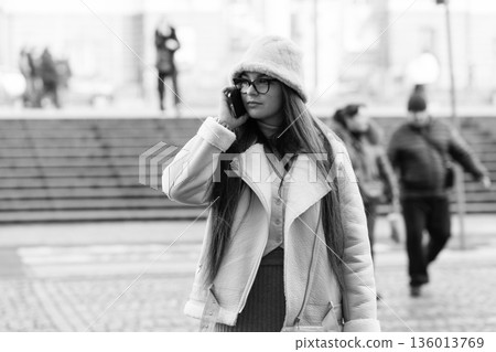 Walking through the bustling city streets, a young woman engages in a lively phone conversation while dressed stylishly for the cool weather Walking through the bustling city streets, a young woman engages in a lively phone conversation while dressed stylishly for the cool weather 136013769
