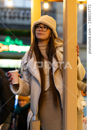 Charming winter evening stroll captures a fashionable woman enjoying a warm beverage amid festive city lights and bustling crowd during twilight hours 136013771