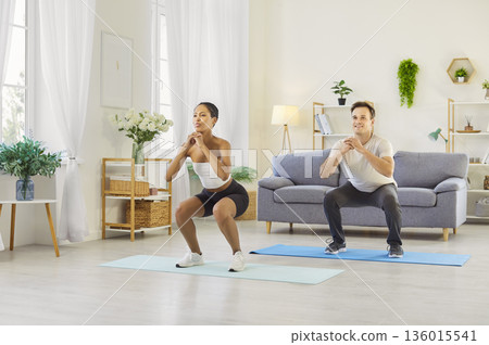 Couple doing squats together at home, building strength and balance in living room Couple doing squats together at home, building strength and balance in living room 136015541
