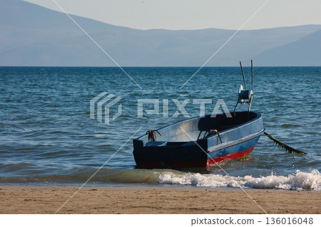Silhouetted fishing boat on the shore of Vlora, Albania, captured on August 24, 2025. The backlit scene shows shimmering sunlight on the sea, gentle waves, and a swimmer in the background, creating a  136016048