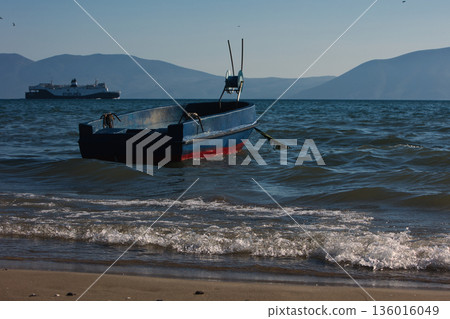 Silhouetted fishing boat on the shore of Vlora, Albania, captured on August 24, 2025. The backlit scene shows shimmering sunlight on the sea, gentle waves, and a swimmer in the background, creating a  136016049