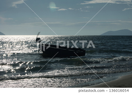 Silhouetted fishing boat on the shore of Vlora, Albania, captured on August 24, 2025. The backlit scene shows shimmering sunlight on the sea, gentle waves, and a swimmer in the background, creating a  136016054