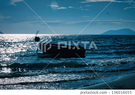 Silhouetted fishing boat on the shore of Vlora, Albania, captured on August 24, 2025. The backlit scene shows shimmering sunlight on the sea, gentle waves, and a swimmer in the background, creating a Silhouetted fishing boat on the shore of Vlora, Albania, captured on August 24, 2025. The backlit scene shows shimmering sunlight on the sea, gentle waves, and a swimmer in the background, creating a 136016055