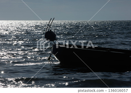 Silhouetted fishing boat on the shore of Vlora, Albania, captured on August 24, 2025. The backlit scene shows shimmering sunlight on the sea, gentle waves, and a swimmer in the background, creating a  136016057