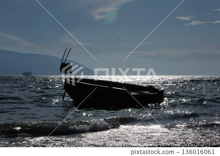 Silhouetted fishing boat on the shore of Vlora, Albania, captured on August 24, 2025. The backlit scene shows shimmering sunlight on the sea, gentle waves, and a swimmer in the background, creating a Silhouetted fishing boat on the shore of Vlora, Albania, captured on August 24, 2025. The backlit scene shows shimmering sunlight on the sea, gentle waves, and a swimmer in the background, creating a 136016061