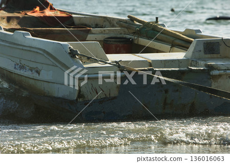 Silhouetted fishing boat on the shore of Vlora, Albania, captured on August 24, 2025. The backlit scene shows shimmering sunlight on the sea, gentle waves, and a swimmer in the background, creating a  136016063