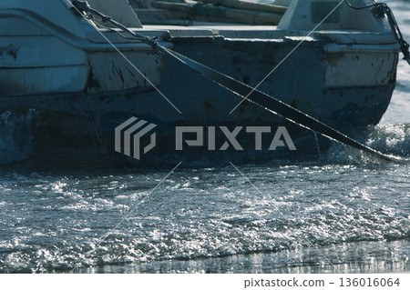 Silhouetted fishing boat on the shore of Vlora, Albania, captured on August 24, 2025. The backlit scene shows shimmering sunlight on the sea, gentle waves, and a swimmer in the background, creating a  136016064