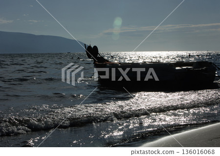 Silhouetted fishing boat on the shore of Vlora, Albania, captured on August 24, 2025. The backlit scene shows shimmering sunlight on the sea, gentle waves, and a swimmer in the background, creating a Silhouetted fishing boat on the shore of Vlora, Albania, captured on August 24, 2025. The backlit scene shows shimmering sunlight on the sea, gentle waves, and a swimmer in the background, creating a 136016068