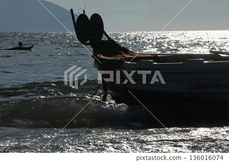 Silhouetted fishing boat on the shore of Vlora, Albania, captured on August 24, 2025. The backlit scene shows shimmering sunlight on the sea, gentle waves, and a swimmer in the background, creating a Silhouetted fishing boat on the shore of Vlora, Albania, captured on August 24, 2025. The backlit scene shows shimmering sunlight on the sea, gentle waves, and a swimmer in the background, creating a 136016074