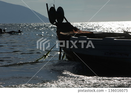 Silhouetted fishing boat on the shore of Vlora, Albania, captured on August 24, 2025. The backlit scene shows shimmering sunlight on the sea, gentle waves, and a swimmer in the background, creating a Silhouetted fishing boat on the shore of Vlora, Albania, captured on August 24, 2025. The backlit scene shows shimmering sunlight on the sea, gentle waves, and a swimmer in the background, creating a 136016075