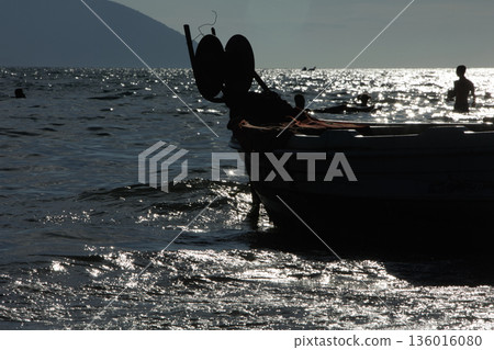 Silhouetted fishing boat on the shore of Vlora, Albania, captured on August 24, 2025. The backlit scene shows shimmering sunlight on the sea, gentle waves, and a swimmer in the background, creating a Silhouetted fishing boat on the shore of Vlora, Albania, captured on August 24, 2025. The backlit scene shows shimmering sunlight on the sea, gentle waves, and a swimmer in the background, creating a 136016080