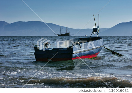 Silhouetted fishing boat on the shore of Vlora, Albania, captured on August 24, 2025. The backlit scene shows shimmering sunlight on the sea, gentle waves, and a swimmer in the background, creating a  136016081