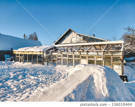 An old wooden greenhouse half covered with deep snow on a sunny winter day. Blue skies overhead and a rural farmhouse in the background creating a peaceful, frosty rural scene. An old wooden greenhouse half covered with deep snow on a sunny winter day. Blue skies overhead and a rural farmhouse in the background creating a peaceful, frosty rural scene. 136018469