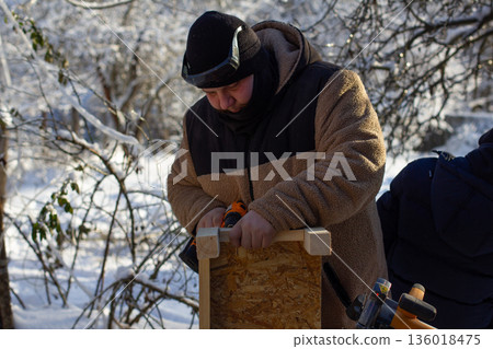 Man in warm winter clothing engaging in a diy project, constructing a wooden birdhouse outdoors during a cold, snowy winter day, symbolizing hobby, craftsmanship, and caring for nature. Man in warm winter clothing engaging in a diy project, constructing a wooden birdhouse outdoors during a cold, snowy winter day, symbolizing hobby, craftsmanship, and caring for nature. 136018475