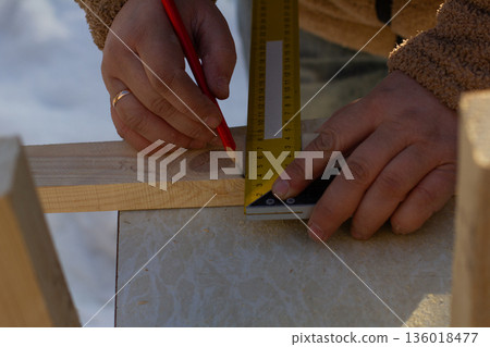 Craftsperson hands accurately measuring a wooden plank with a ruler and marking it with a red pencil, demonstrating precision in woodworking and construction. 136018477