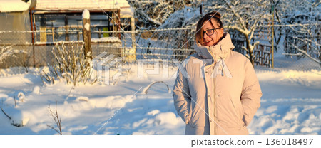 A woman stands in a snowy courtyard wearing a warm down jacket and sunglasses, smiling and looking away. On a bright, cold winter day, the woman enjoys the peaceful scenery outside. 136018497