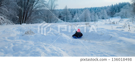 Child wearing a pink hat having fun sledding down a snowy slope, surrounded by a serene winter landscape with frost-covered trees creating a peaceful and playful childhood scene. 136018498