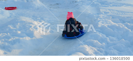 A girl in a pink hat and winter coat rides down a snowy slope on a blue sled, shot from behind. The child laughs and races down the icy hill against the backdrop of a bright, crisp winter landscape. A girl in a pink hat and winter coat rides down a snowy slope on a blue sled, shot from behind. The child laughs and races down the icy hill against the backdrop of a bright, crisp winter landscape. 136018500