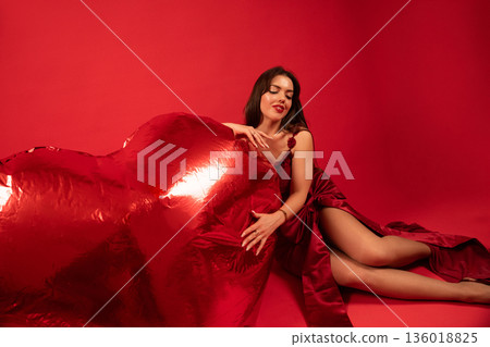 Woman sits with large red heart while wearing a red dress in a studio setting for Valentines Day celebration 136018825