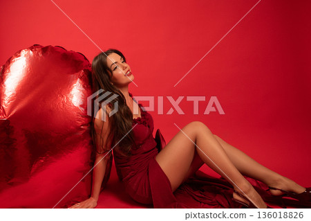 Woman in red dress poses next to heart decoration for Valentines Day celebration in indoor setting Woman in red dress poses next to heart decoration for Valentines Day celebration in indoor setting 136018826
