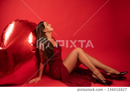 Beautiful woman sitting by a large red heart on Valentines Day in a studio Beautiful woman sitting by a large red heart on Valentines Day in a studio 136018827
