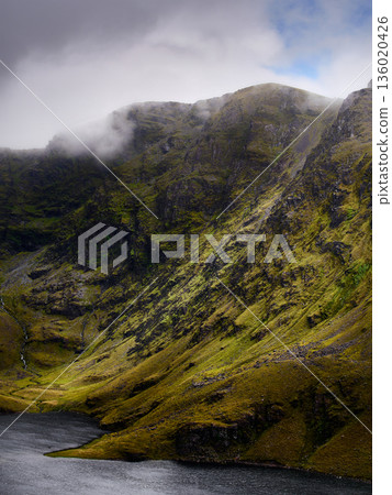 A serene lake sits in a deep valley surrounded by green slopes and a cascade falling from the mountains. Clouds hang low over the mountain peaks. 136020426