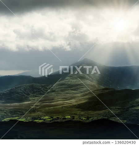 A moody landscape bathed in golden rays, revealing forested terrain and distant peaks. 136020430