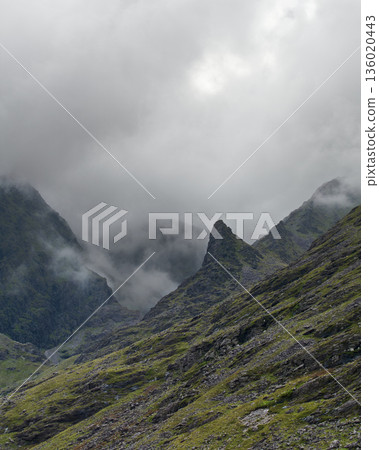 Cloud-covered mountain landscape showcasing steep rocky slopes and jagged peaks on a dim day. Cloud-covered mountain landscape showcasing steep rocky slopes and jagged peaks on a dim day. 136020443