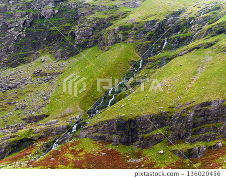 Green rocky mountain slopes feature numerous small waterfalls cascading downwards, creating a vibrant natural scene. 136020456