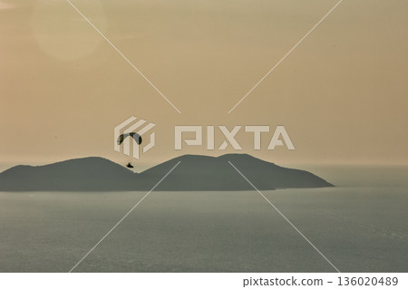 Mountain range rising above a calm blue sea under a clear sky, forming a minimalistic coastal landscape. Serene natural scenery captured on August  2025. 136020489
