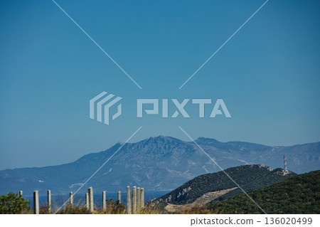 Mountain range rising above a calm blue sea under a clear sky, forming a minimalistic coastal landscape. Serene natural scenery captured on August  2025. 136020499