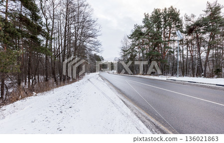 A quiet winter scene features a snow-dusted road winding through a forest 136021863