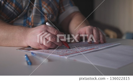 A man sits at a table writing notes with a red pen in his right hand. Blue pens and loose papers are nearby while he focuses on his work during the afternoon light. A man sits at a table writing notes with a red pen in his right hand. Blue pens and loose papers are nearby while he focuses on his work during the afternoon light. 136022815