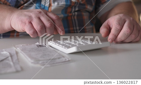 A person sits at a table pressing buttons on a number pad. Receipts and a check lie on the table. This scene happens indoors in a well-lit area during the day. A person sits at a table pressing buttons on a number pad. Receipts and a check lie on the table. This scene happens indoors in a well-lit area during the day. 136022817