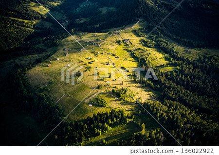 Aerial view of sunlit valley, dotted with small houses and winding roads, surrounded by dense forests. Golden light casts long shadows. Vibrant landscape stretches out, merging into forested hills. 136022922