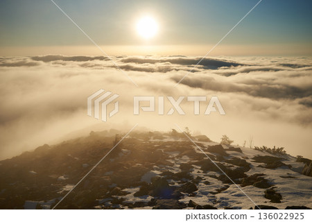 Sunrise over snow-covered mountain peak. Warm golden light of rising sun bathes clouds in soft hues of orange and pink. Snow and scattered rocks in foreground add texture and depth to scene. Sunrise over snow-covered mountain peak. Warm golden light of rising sun bathes clouds in soft hues of orange and pink. Snow and scattered rocks in foreground add texture and depth to scene. 136022925