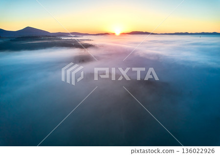 Stunning aerial view of sunrise over mountainous landscape, with thick fog blanketing valleys and hills. Sun's golden rays pierce through mist, casting serene glow over the entire scene. 136022926