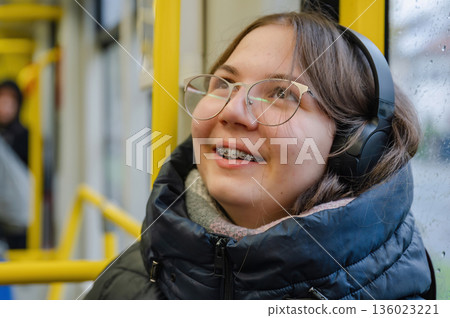 Smiling teenager girl with braces and headphones in tram during rainy weather 136023221