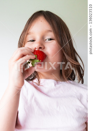 Caucasian young girl enjoying fresh strawberry in bright white setting 136023510