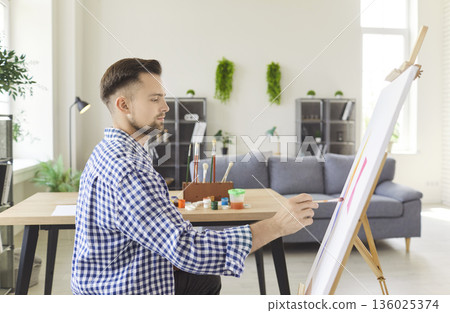 young man artist sitting in front of easel at home workshop, making picture young man artist sitting in front of easel at home workshop, making picture 136025374