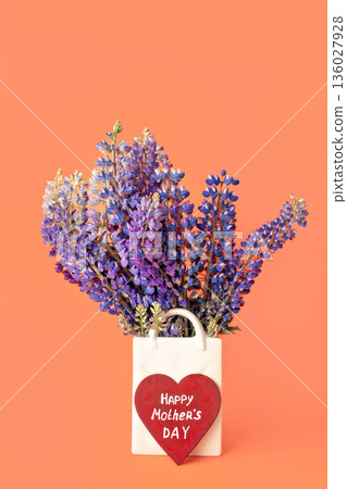 Bouquet of fresh lupine flowers in white vase shaped like bag and wooden red heart with white handwritten inscription happy Mother's day on peach color background, vertical Bouquet of fresh lupine flowers in white vase shaped like bag and wooden red heart with white handwritten inscription happy Mother's day on peach color background, vertical 136027928