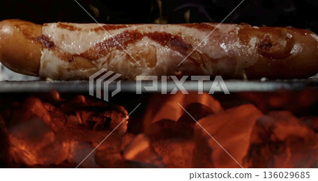 Bacon wrapped hot dog cooking on grill with burning charcoal flames in background during evening barbecue event Bacon wrapped hot dog cooking on grill with burning charcoal flames in background during evening barbecue event 136029685