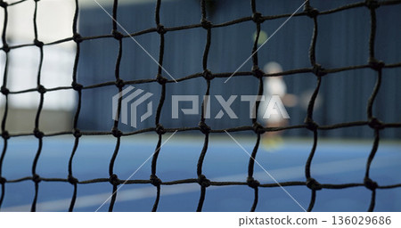 Tennis player practices serve on blue court in indoor facility near a net during afternoon session 136029686